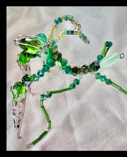 A handmade beaded insect ornament, resembling a mantis, crafted with faceted green and light blue beads, clear glass elements, and silver wire on a textured white fabric background.