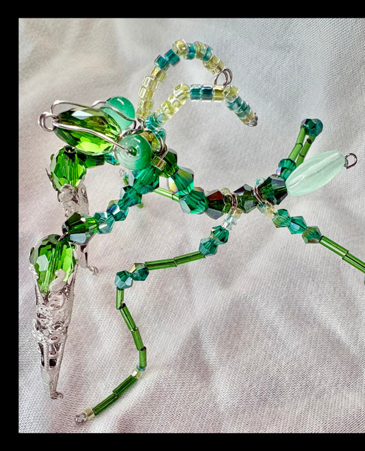 A handmade beaded insect ornament, resembling a mantis, crafted with faceted green and light blue beads, clear glass elements, and silver wire on a textured white fabric background.